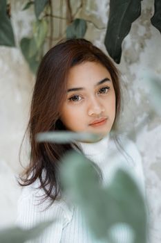 一位身着白色高领毛衣的黑发年轻女子注视着镜头，周围的树叶一片模糊