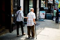 An elderly man with a cane is assisted by a younger companion walking along a busy New York City street.