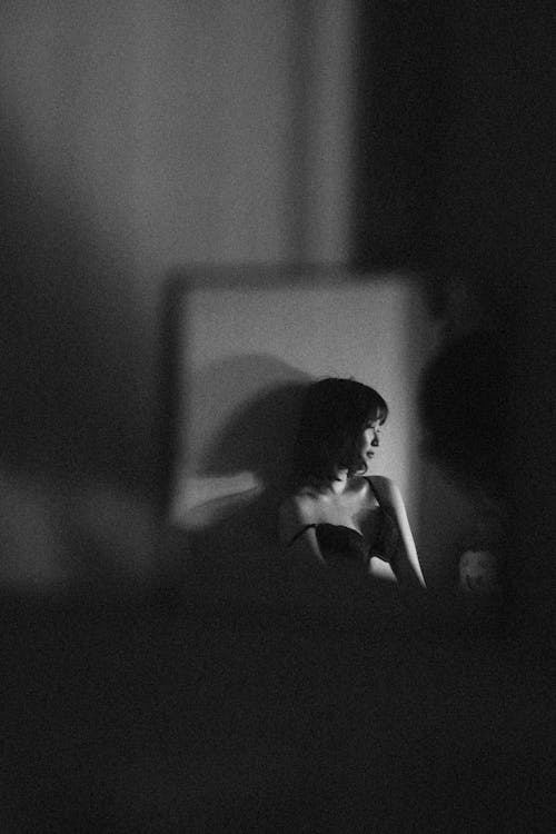 Through window frame black and white of unrecognizable distant female in bra leaning and casting shadow on wall in dark room while looking away