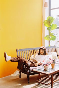 Full body of young Asian female in cap lying on wooden bench with magnifier in hand near table with potted plants in room with yellow wall