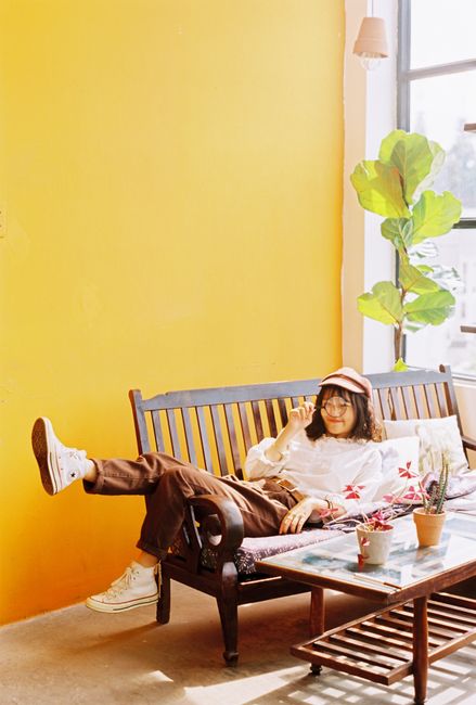 Full body of young Asian female in cap lying on wooden bench with magnifier in hand near table with potted plants in room with yellow wall