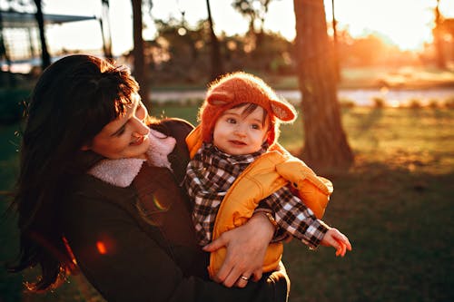 Free Cheerful mother smiling and holding baby in funny hat and bright vest in garden Stock Photo