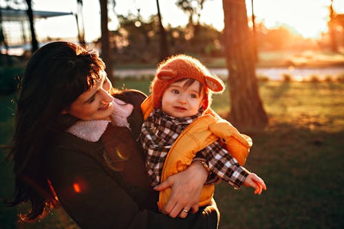 Cheerful mother smiling and holding baby in funny hat and bright vest in garden