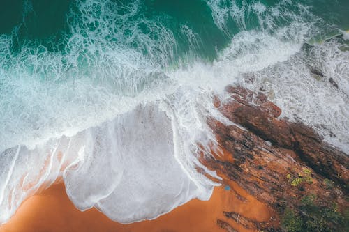 Free Aerial view of foamy waves of ocean washing sandy beach with rock in sunny day Stock Photo
