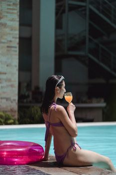 Back view of female wearing bikini drinking fresh fruit juice while sitting on poolside in sunny day