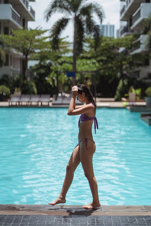 Side view full body attractive fit female in bikini walking on wet poolside in tropical resort on hot sunny day
