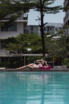 Full body slender gorgeous female in purple swimwear lying graciously on poolside in sunny tropical resort
