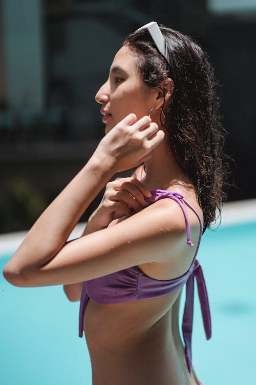 Side view attractive fit female in violet swimwear standing in pool and touching face tenderly while chilling in sunny resort