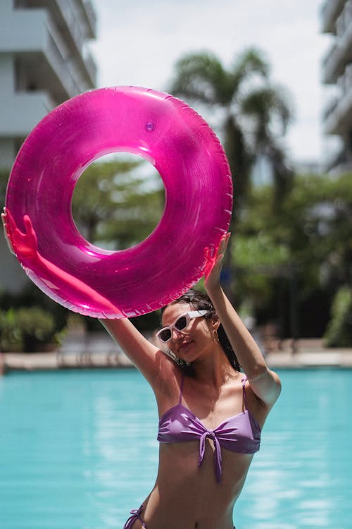 Content slim female in purple swimwear standing on sunny poolside and raising pink inflatable swimming ring