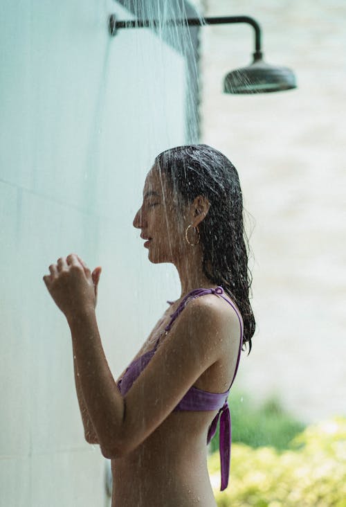 Slim young lady in swimwear standing under shower on sunny day