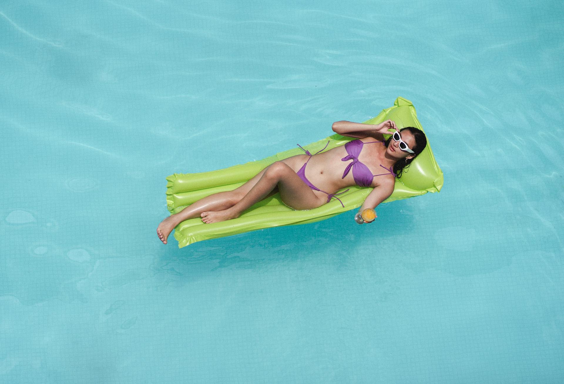 From above of delighted young slim tanned lady in bikini and sunglasses relaxing with orange juice in swimming pool