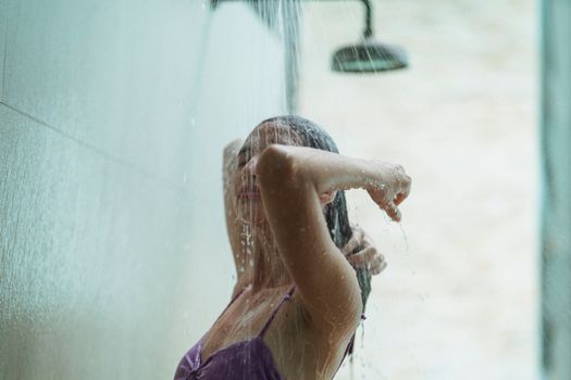 Attractive slim woman with long dark hair wearing violet swimwear rinsing off in warm shower after swimming