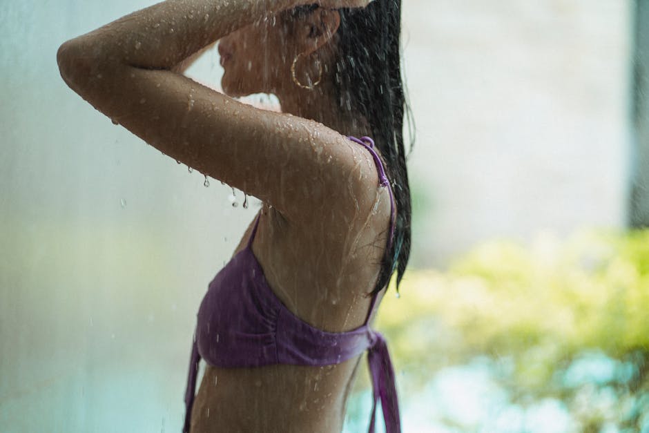Side view of crop sensual slim female wearing lilac swimsuit standing in shower and raising hands to rinse off hair on blurred background