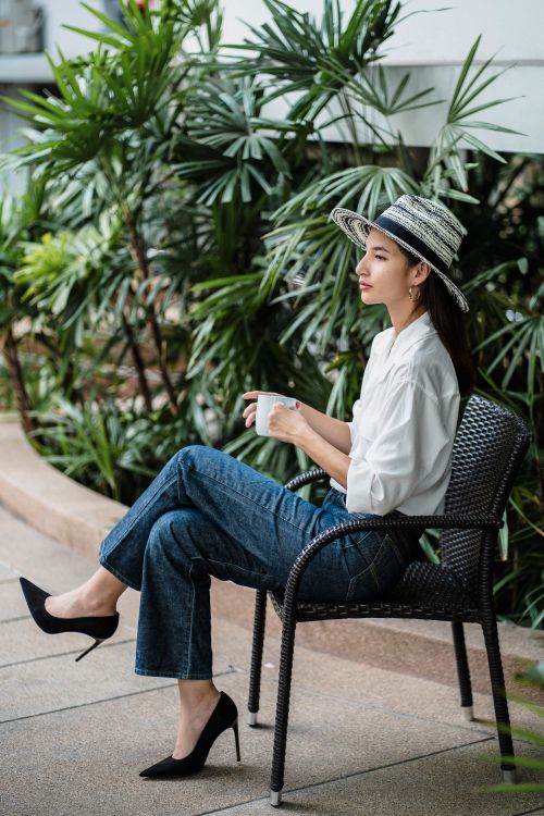 Free Side view of ethnic female in high heels and stylish denim with white shirt resting with tea in armchair next to palm trees Stock Photo