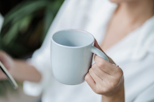 Free From above of crop unrecognizable lady in white blouse enjoying delicious beverage in ceramic cup while reading book Stock Photo