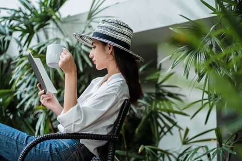 Free Side view of stylish young lady in trendy clothes sitting in garden on black chair with book and ceramic mug with hot coffee Stock Photo