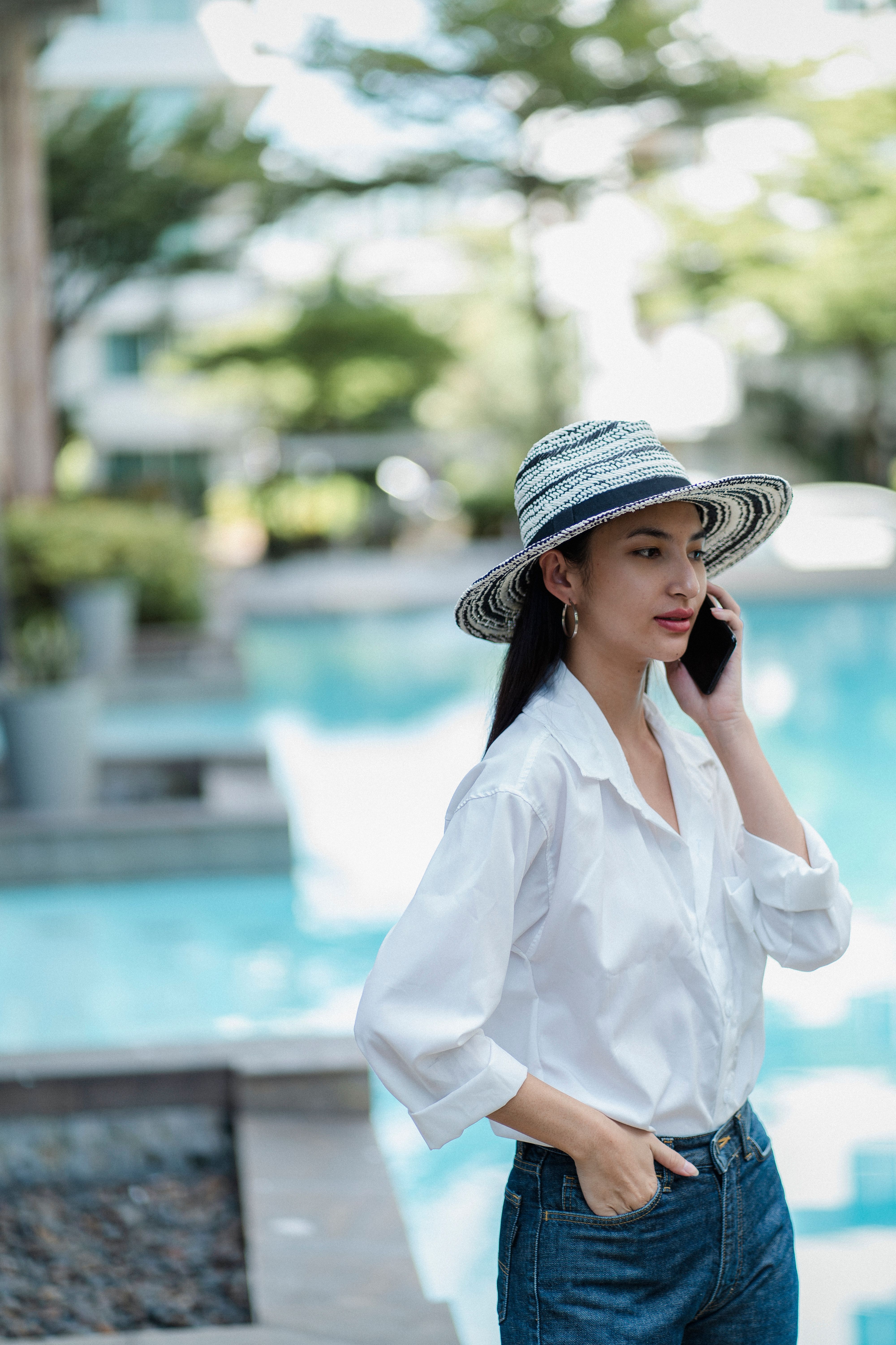 Pensive stylish lady in hat standing near modern pool and speaking on smartphone on summer day