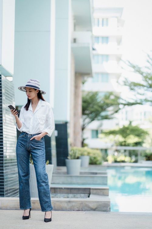 Free Full body of calm young lady in hat and stylish outfit using mobile and standing near modern pool on summer day Stock Photo