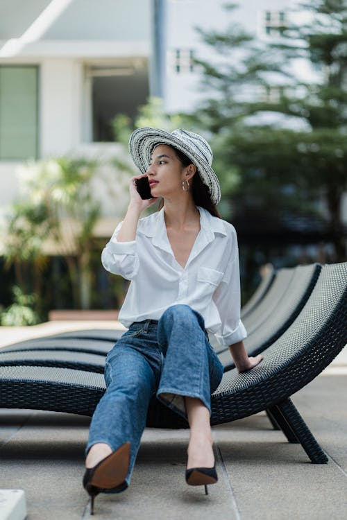 Full body of confident young Asian lady in stylish outfit and hat sitting on sunbed in courtyard and having phone conversation