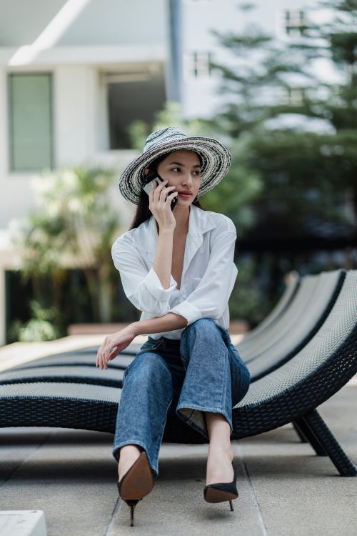 Free Full length of stylish young ethnic female in trendy jeans and hat sitting on lounger and looking away while talking on mobile phone on sunny day Stock Photo