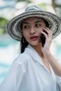Side view of gorgeous young Asian female in trendy hat and white shirt talking on mobile phone and looking over shoulder near outdoor pool