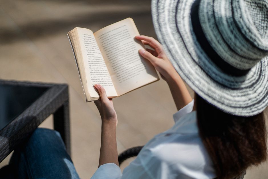 From above of crop anonymous female in stylish outfit and hat reading interesting book while relaxing in outdoor cafe