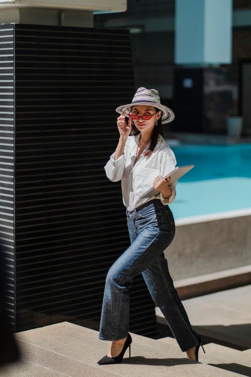Free Full body of self assured young Asian female freelancer in trendy outfit and hat with laptop in hand walking upstairs near outdoor swimming pool and talking on phone Stock Photo