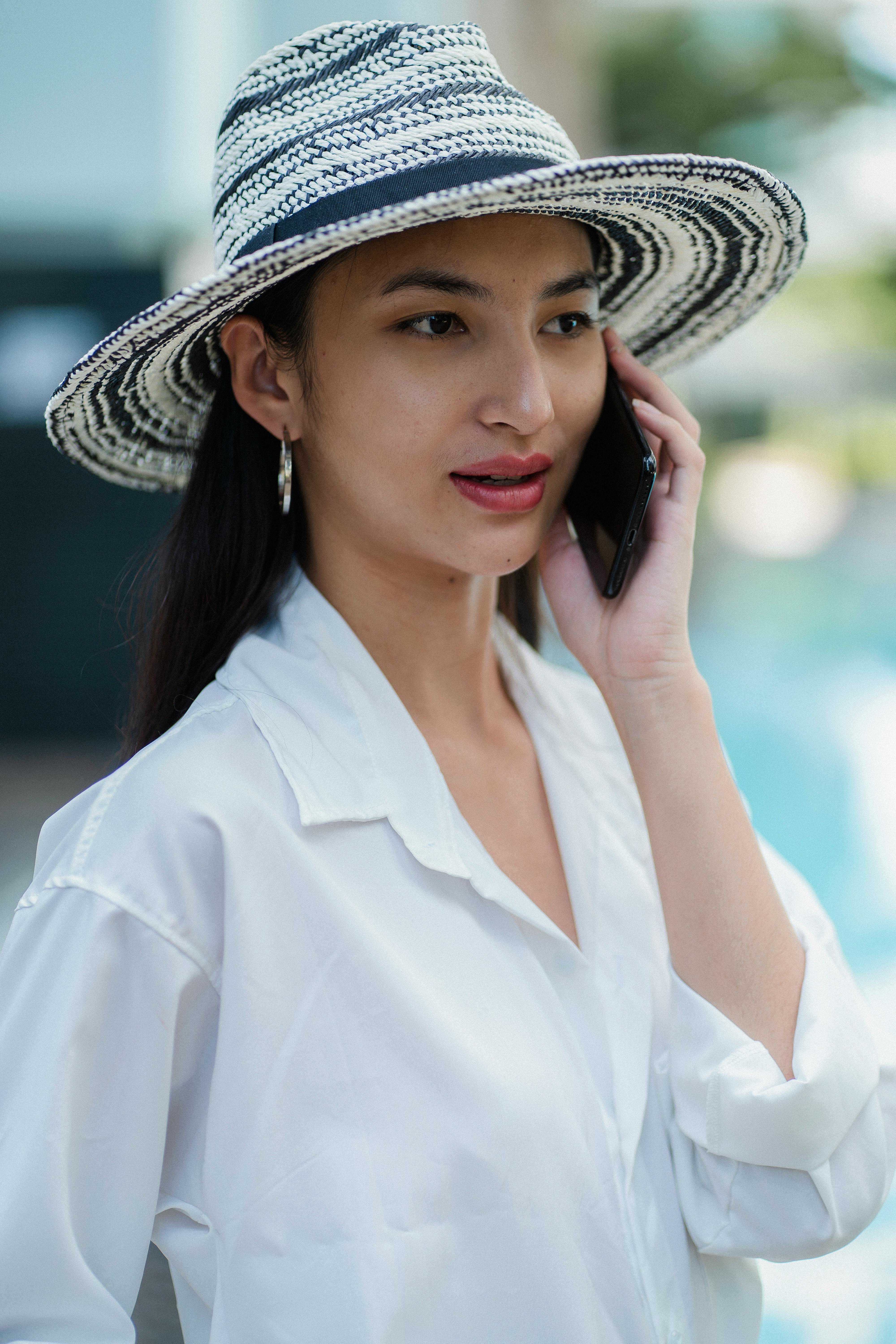 Concentrated young Asian lady in white shirt and straw hat talking on cellphone and looking away on sunny day