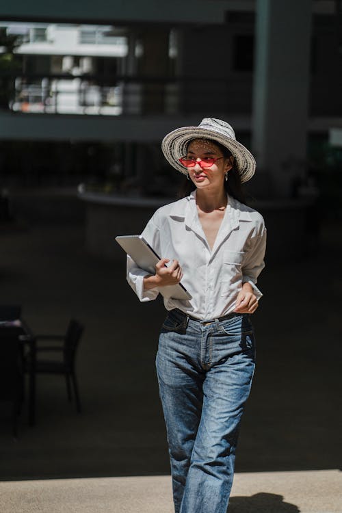 Stylish young ethnic female entrepreneur in trendy hat and sunglasses walking on street with laptop in hand and looking away on sunny day