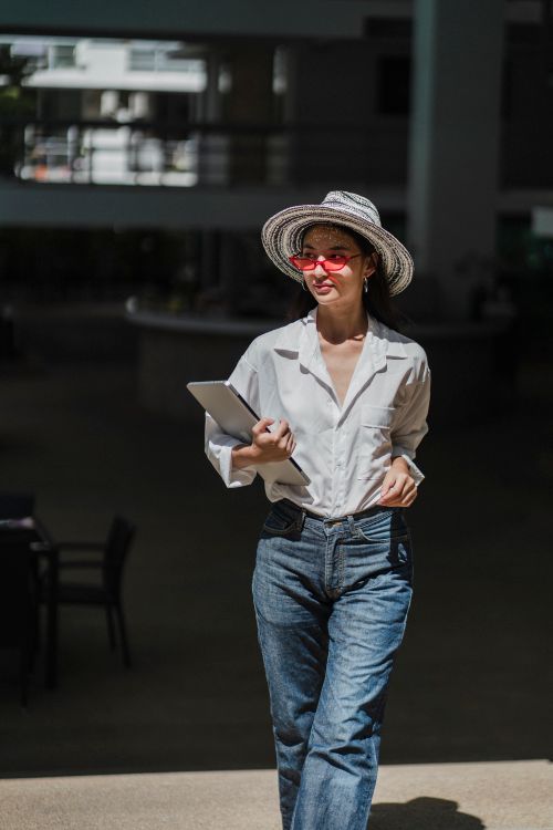 Free Stylish young ethnic female entrepreneur in trendy hat and sunglasses walking on street with laptop in hand and looking away on sunny day Stock Photo
