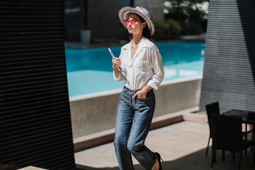 Free Self assured young ethnic lady in elegant outfit and sunglasses walking near outdoor swimming pool with laptop in hand during summer vacation Stock Photo