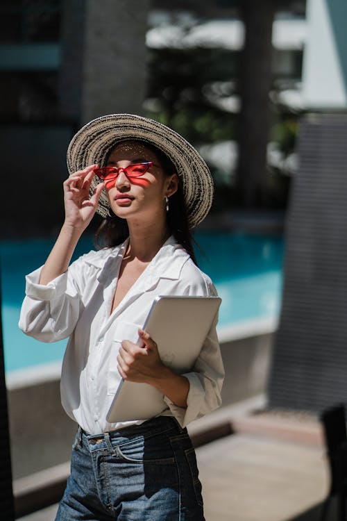Free Self esteem young Asian female freelancer in trendy clothes and hat adjusting sunglasses while standing near swimming pool with laptop in hand Stock Photo