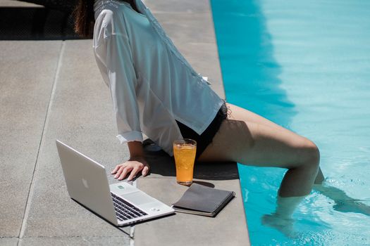 From above side view of crop unrecognizable female traveler sitting on pool border with legs in water while relaxing after remote job on laptop