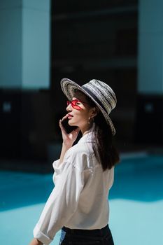 Side view of young fashionable Asian lady with dark hair in straw hat and sunglasses having phone call while standing near outdoor swimming pool on sunny day