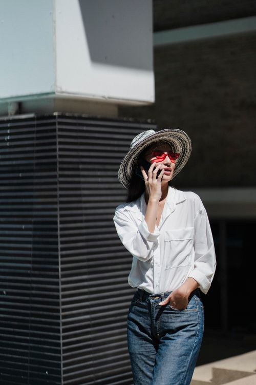 Free Elegant young ethnic female in trendy hat and white shirt walking near modern building with hand in pocket and talking on smartphone Stock Photo