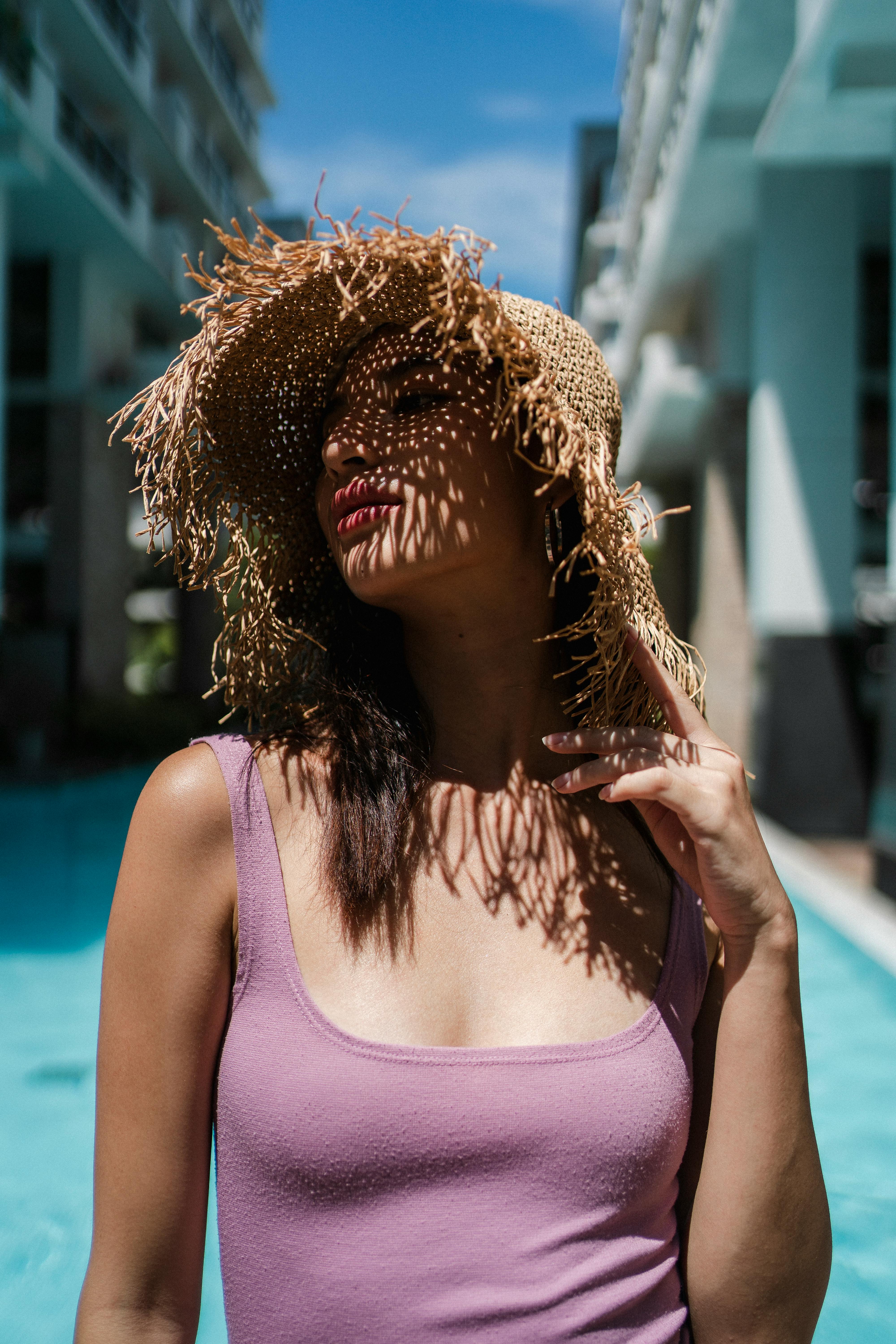 Alluring young ethnic female traveler in top and straw hat relaxing at poolside and looking away during summer holidays in modern hotel