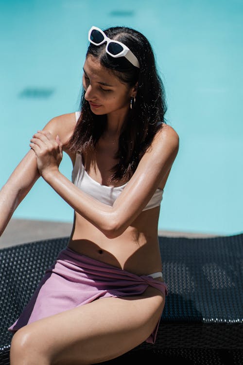 Free From above of young female wearing bikini and wrapped in towel sitting on poolside and spreading sunscreen lotion on body Stock Photo