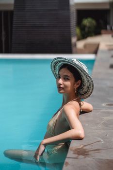 Side view of confident young slim Asian female traveler in stylish swimwear and sunhat leaning on border while resting in pool on sunny day