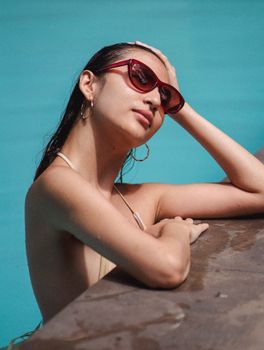 Side view of feminine young ethnic woman with long dark hair in swimwear and sunglasses touching head while relaxing in outdoor pool on sunny summer day