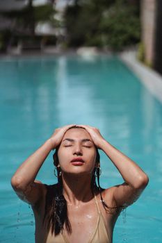 Sensual young Asian female in bikini touching long wet hair with closed eyes while getting out of pool after swimming on sunny day