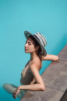 From above side view of self assured young ethnic female model in stylish hat and bikini recreating in swimming pool during summer holidays