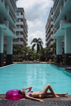 Side view of unrecognizable gorgeous female tourist in swimwear and sunglasses sunbathing lying on edge of pool in modern tropical resort
