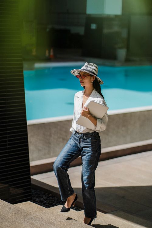 Free Full body of serious young Asian female freelancer in stylish outfit and hat walking upstairs near outdoor swimming pool with modern laptop in hand Stock Photo