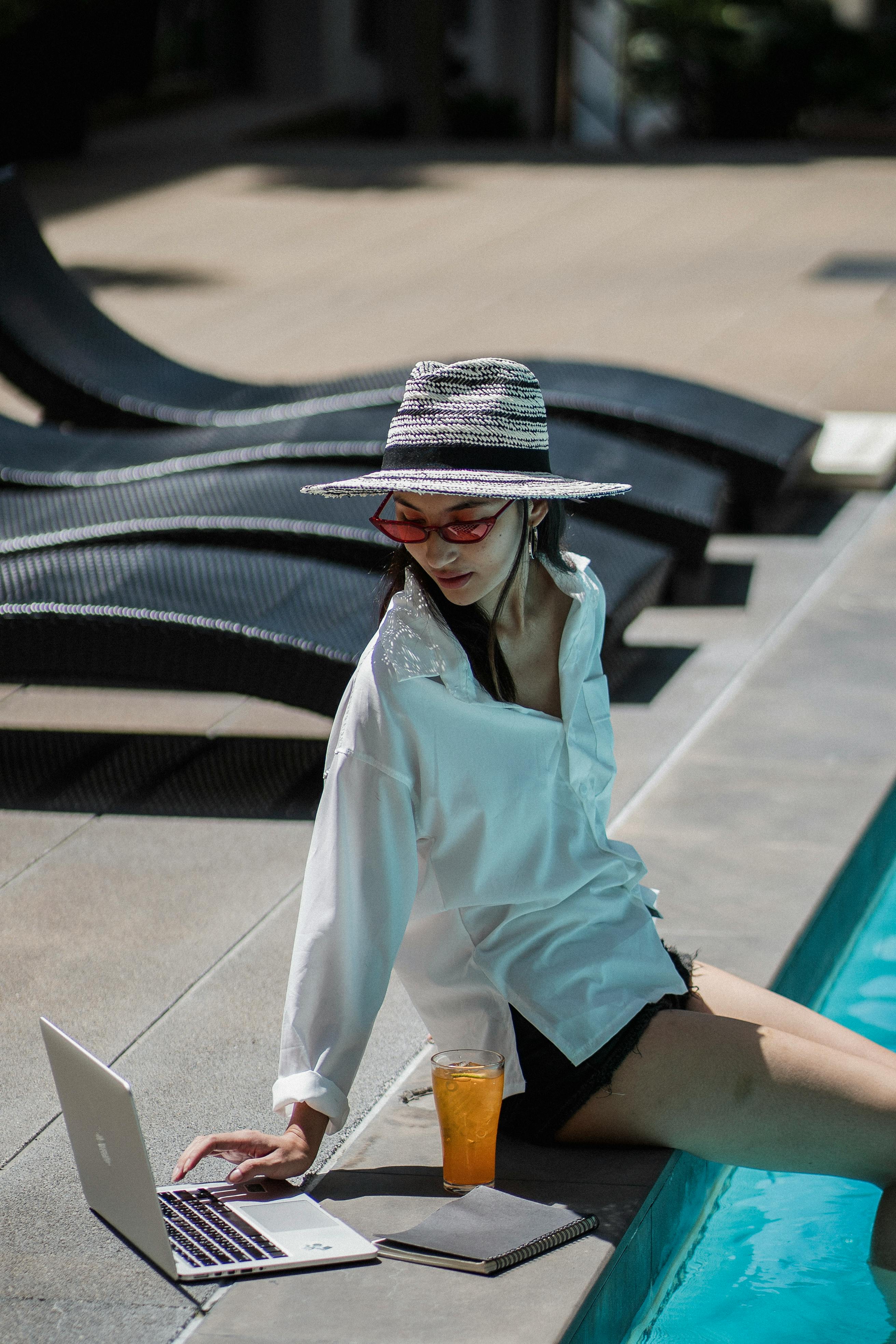 Free Side view of young confident Asian female freelancer in stylish hat and white shirt sitting at poolside and working on laptop during summer vacation Stock Photo