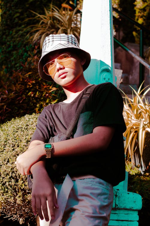 Confident young Asian male wearing hat and sunshades looking at camera while standing near green bushes on sunny street in city