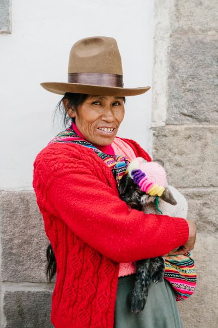 Smiling Peruvian woman in traditional attire holds a lamb with colorful attire against a stone wall.