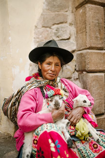 A woman in vibrant traditional clothing holds two goats in a rustic Peruvian setting.