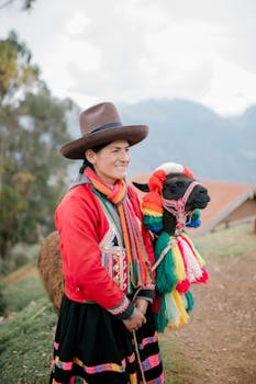 A smiling Peruvian woman in traditional clothing stands with a colorfully adorned llama in the Andes.