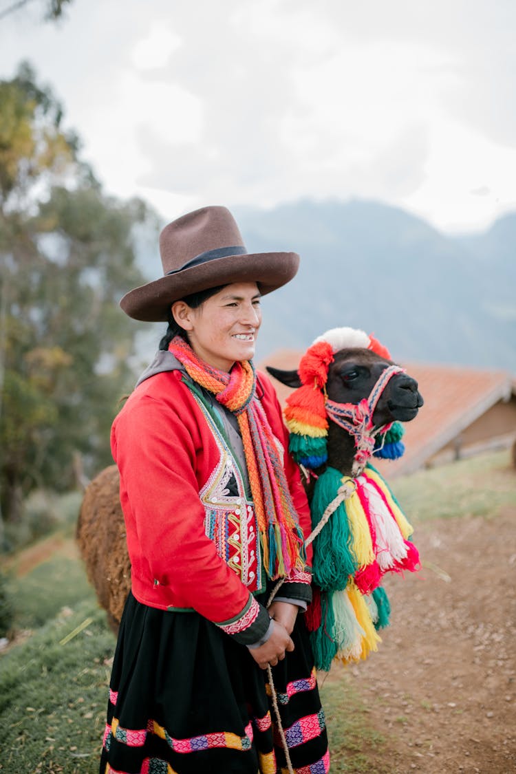 Positive Ethnic Woman With Cute Lama Standing In Peruvian Village On Cloudy Day