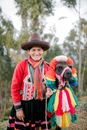 Positive adult native Peruvian female in national dress and hat smiling while standing near trees with cute lama decorated with colorful traditional tassels